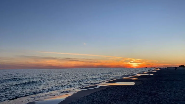 The sun sets on the horizon over calm waves rolling onto a wide sandy beach under an orange and blue sky