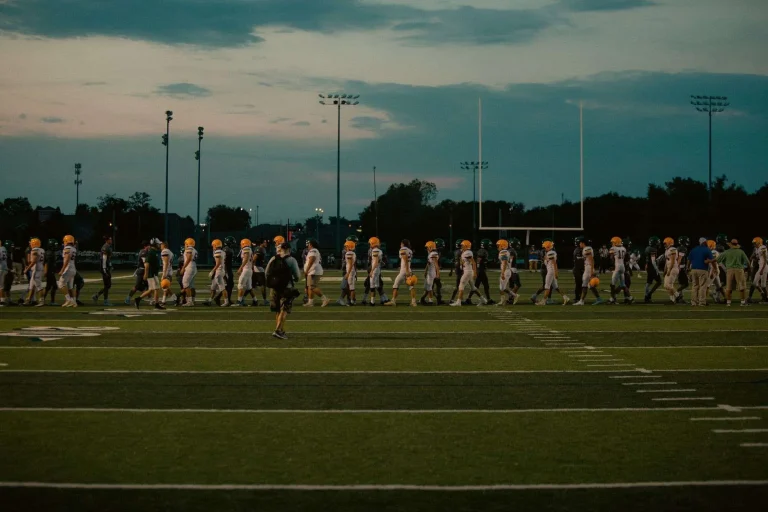 High school football players and coaching staff walking across a field at dusk with stadium lights and a goalpost in the background