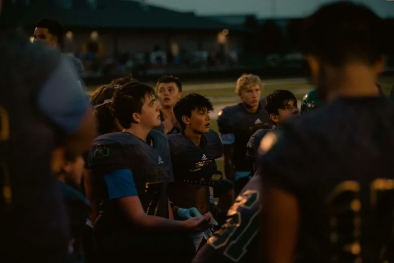 Young football players in dark jerseys gathered in a huddle on a field at dusk