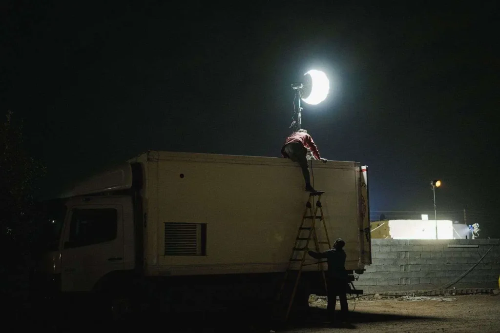 Film crew member setting up lighting equipment atop a production truck at a nighttime outdoor movie set