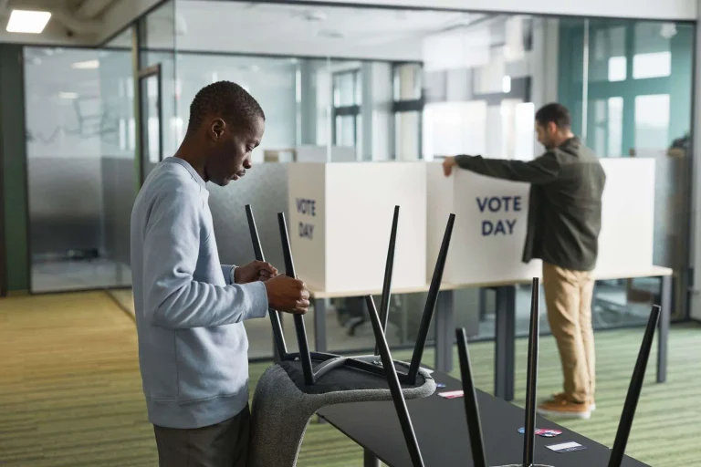 Two people setting up privacy voting booths labeled "Vote Day" in an office-like space, with chairs stacked on a table