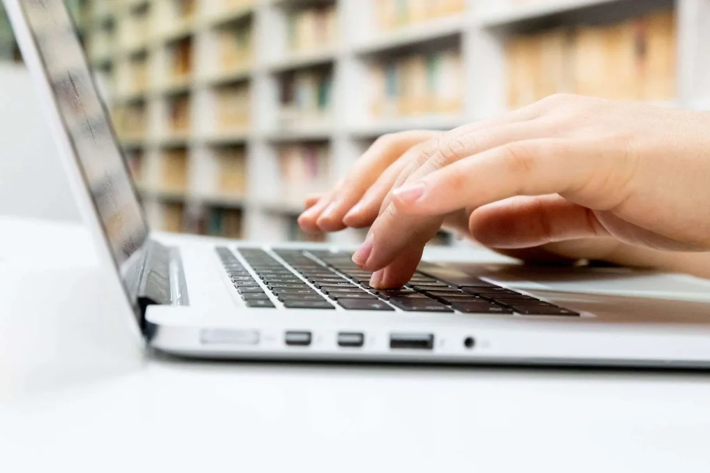 Close-up of hands typing on a laptop keyboard, with bookshelves in the background