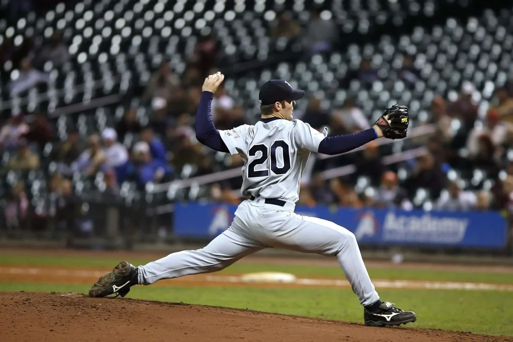 A baseball pitcher in mid-windup delivers a pitch during a night game, with stadium lights and stands in the background
