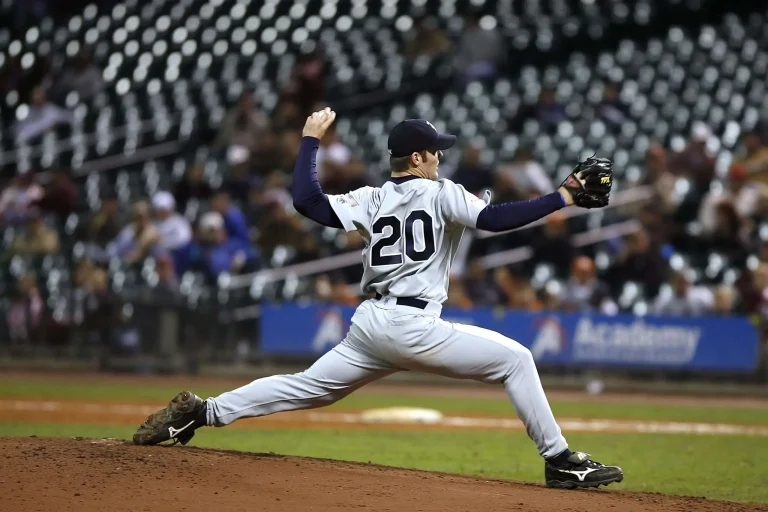 A baseball pitcher in mid-windup delivers a pitch during a night game, with stadium lights and stands in the background