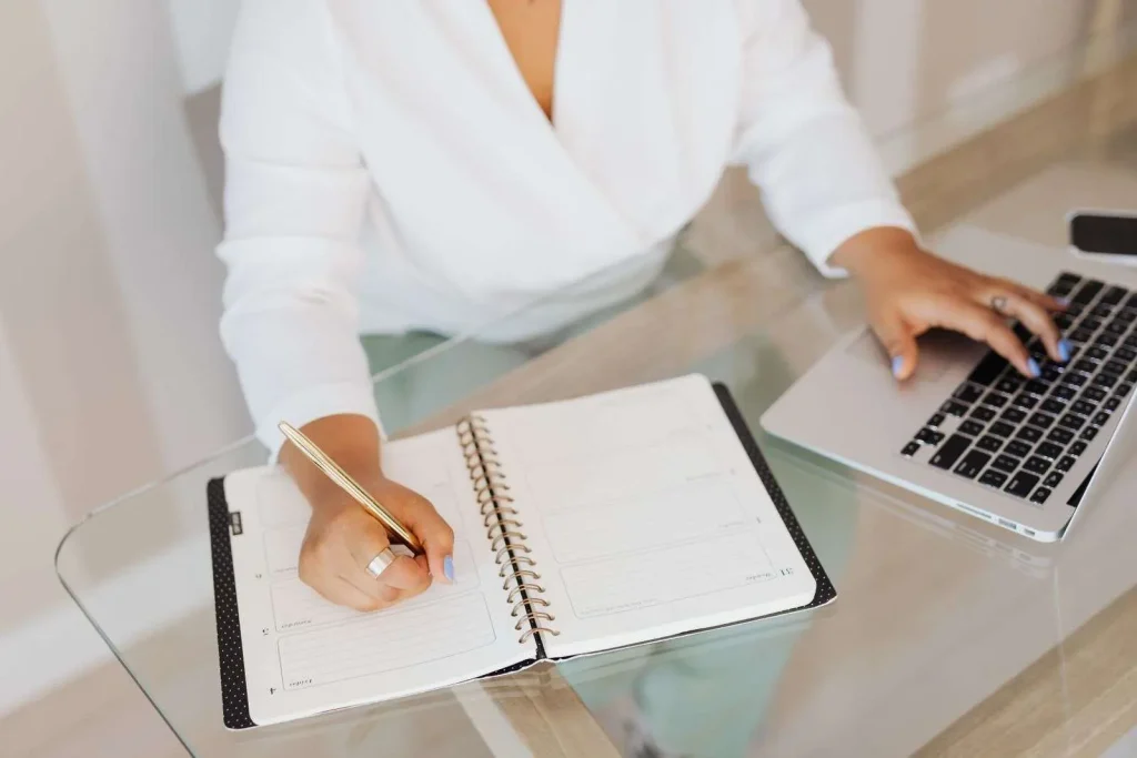 A person writes by hand in a notebook while typing on a laptop at a desk