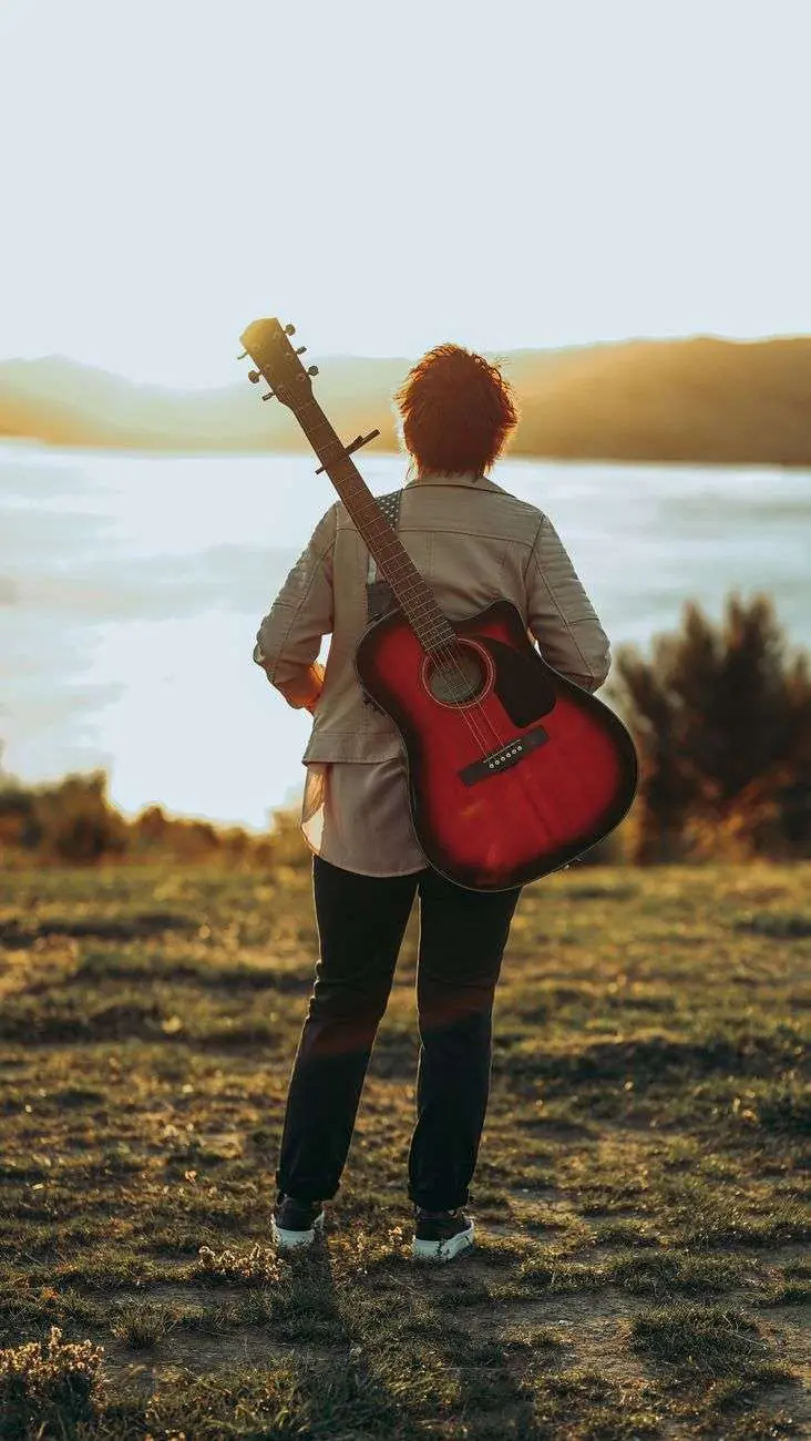 A person seen from behind, guitar slung over one shoulder, standing and looking out over a body of water at sunset