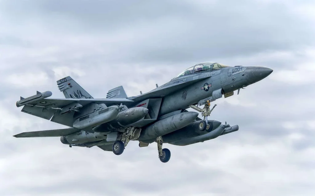 A U.S. Navy F/A-18 Super Hornet fighter jet in flight with landing gear extended, against a cloudy sky.
