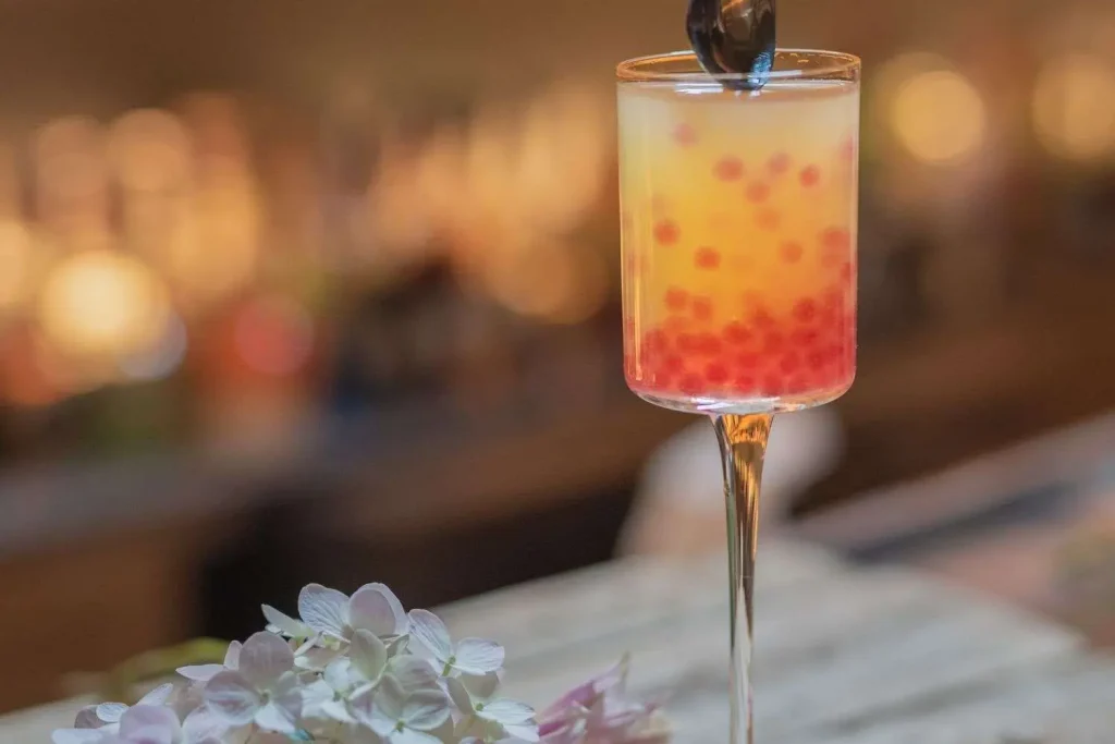 A colorful layered cocktail with red garnish in a stemmed glass, with flowers in the foreground and a dimly lit bar blurred behind