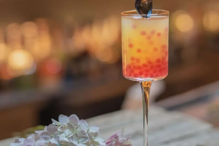 A colorful layered cocktail with red garnish in a stemmed glass, with flowers in the foreground and a dimly lit bar blurred behind