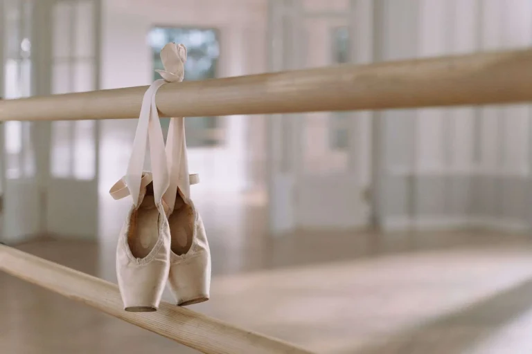 A pair of pink satin pointe shoes hanging from a wooden barre in a bright dance studio