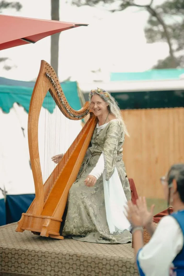 A woman in a medieval-style gown plays a large wooden harp on an outdoor stage at a Renaissance-themed festival, with tents and trees in the background.