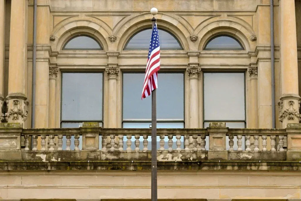 An American flag on a flagpole in front of an ornate stone government building with arched windows and a balustrade balcony