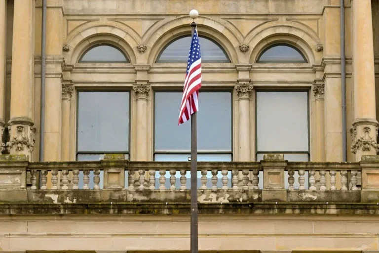 An American flag on a flagpole in front of an ornate stone government building with arched windows and a balustrade balcony