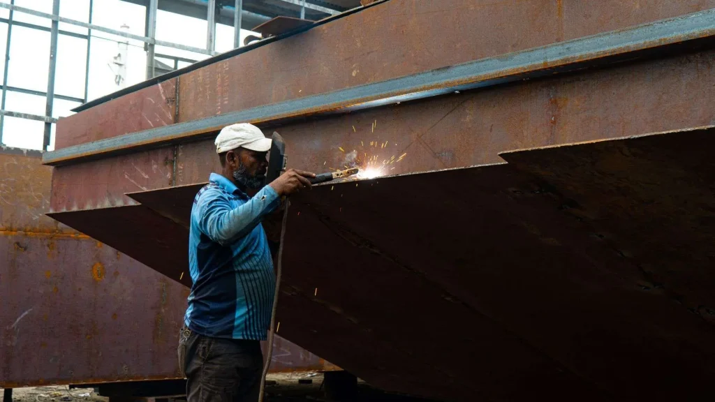 A welder works on a large steel structure, with sparks flying from the weld