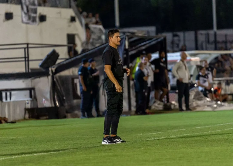 A soccer coach in dark training gear stands on the touchline watching a match under stadium lights