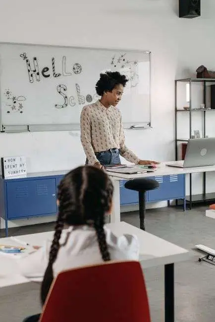 A teacher stands at a classroom whiteboard reading Hello School while a student sits at a desk nearby