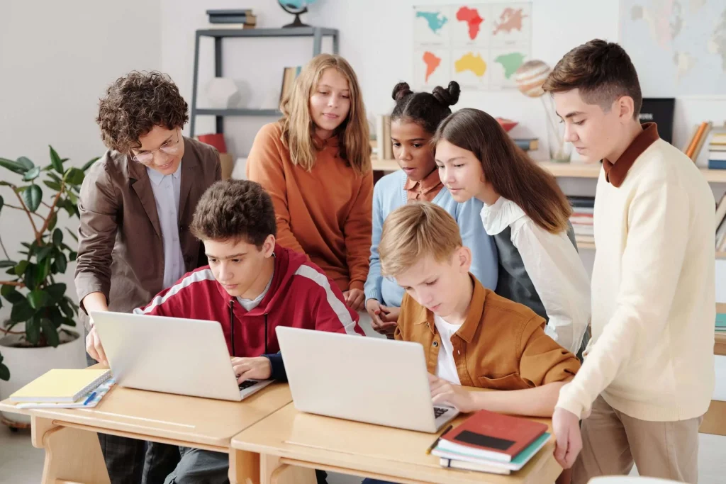 Group of teenage students gathered around laptops with an adult mentor looking on in a classroom