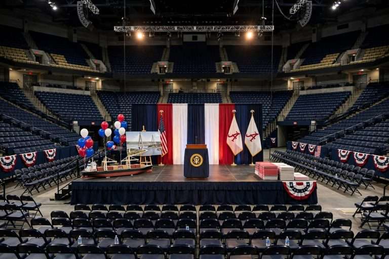 Podium with American flags on a stage inside a crowded college basketball arena lit for an evening political rally