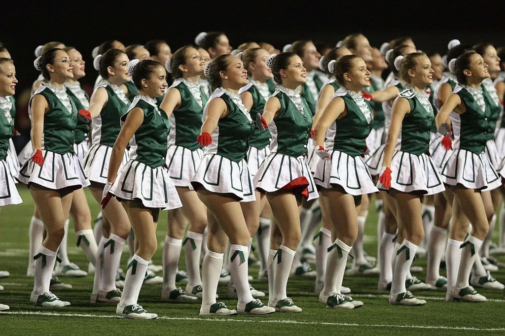 Members of a dance or drill team perform in matching green and white uniforms on a football field at night