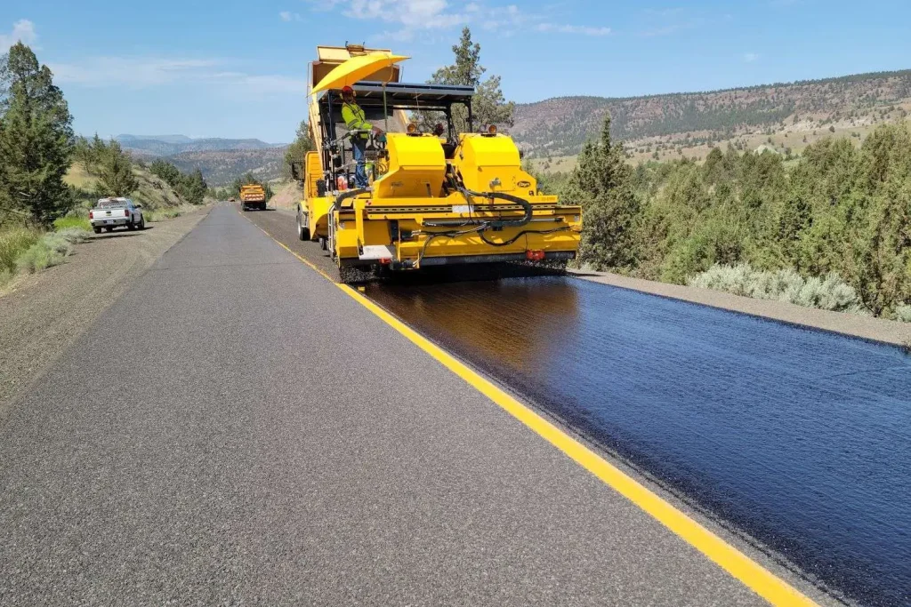 Yellow asphalt paving machine laying fresh blacktop on a two-lane rural highway, with a work truck nearby