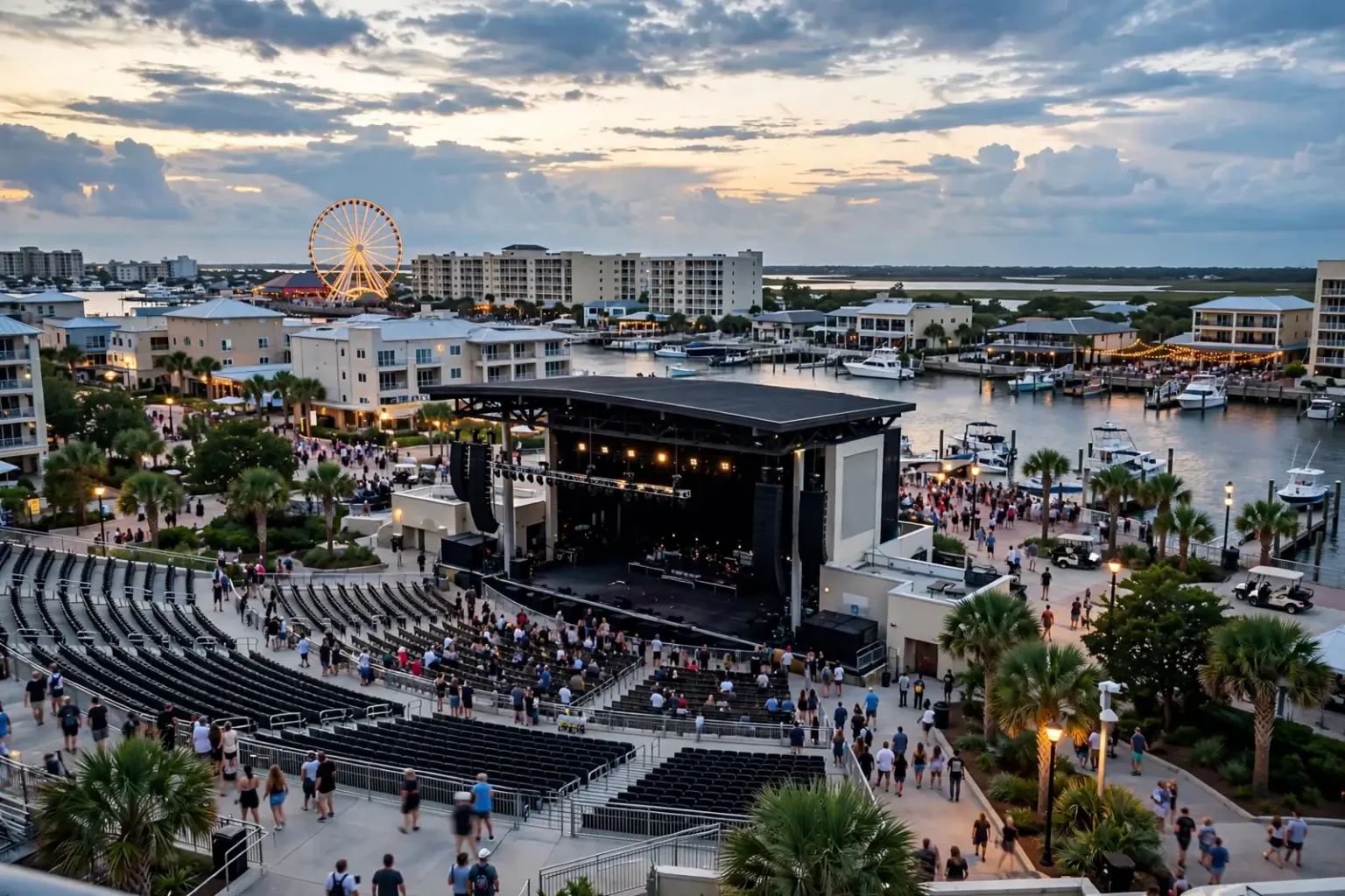 Fans filling the seats of a waterfront amphitheater beside a marina and Ferris wheel at dusk, where Matchbox Twenty will play in Orange Beach