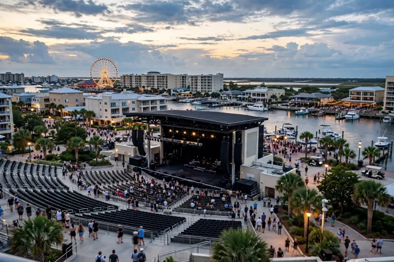 Fans filling the seats of a waterfront amphitheater beside a marina and Ferris wheel at dusk, where Matchbox Twenty will play in Orange Beach
